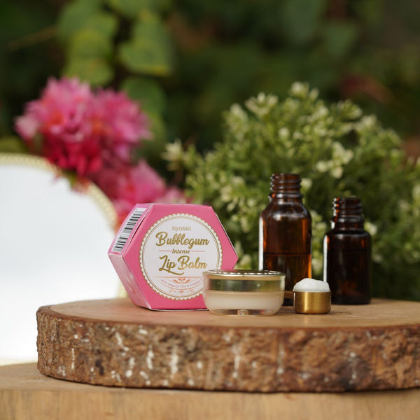 Lip balm, small bottles, and a candle on a wooden surface with flowers in the background
