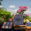 Two makeup palettes with a brand logo on a wooden stand outdoors with flowers and plants.